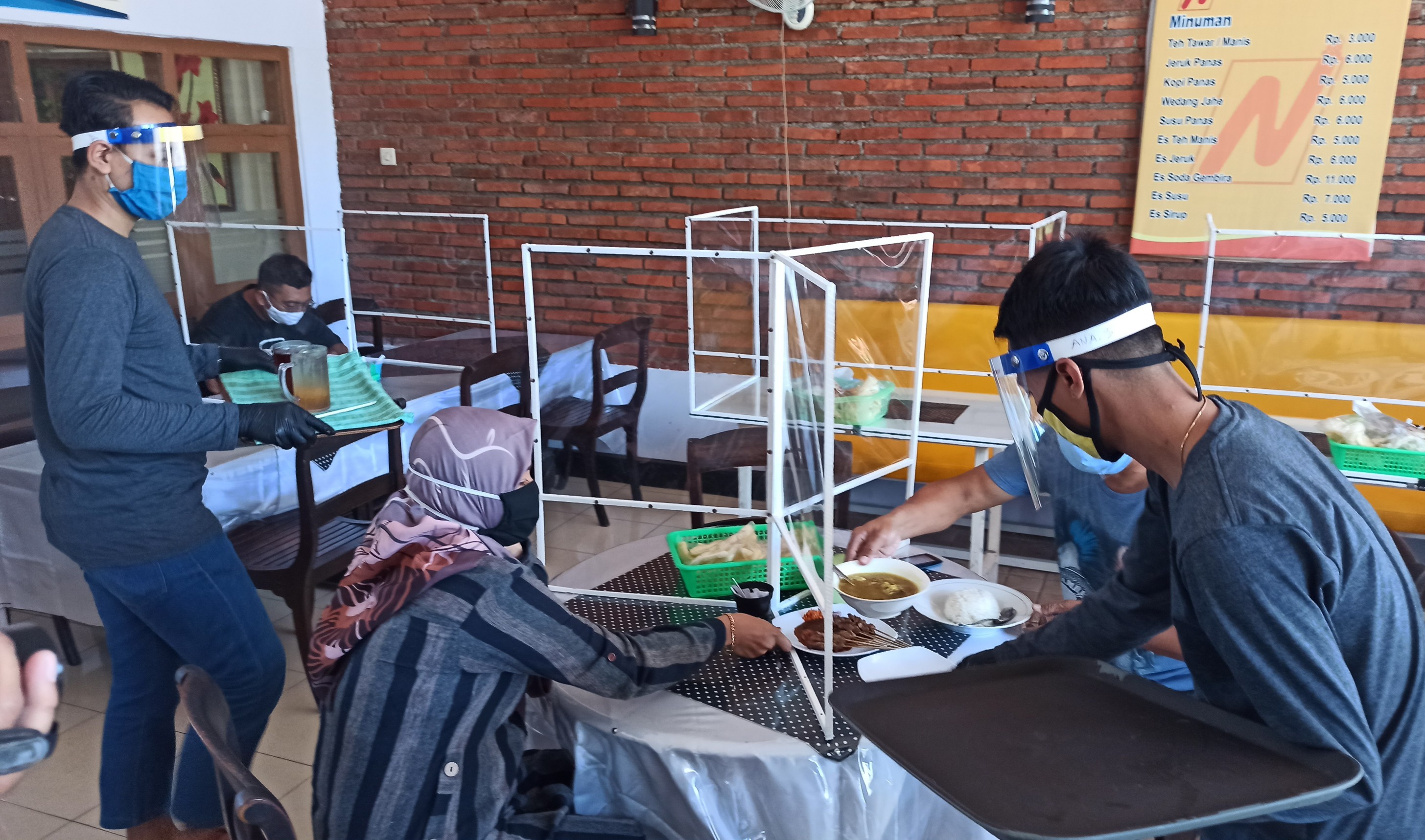 Pelayan Warung Nikmat mengenakan masker, face shield dan sarung tangan sedang mengantar pesanan pengunjung. Pada meja pengunjung tampak pembatas antar pengunjung yang terbuat dari plastik (foto: Hujaini/ngopibareng.id)