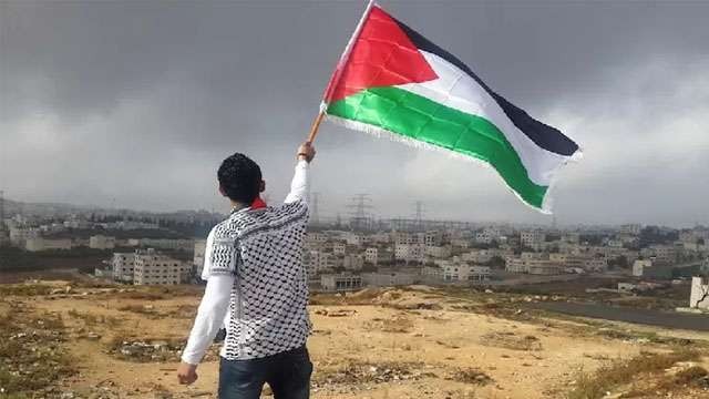 Ilustrasi tulisan; Mengapa  Tuhan Tidak Segera  Hancurkan Israel? Seorang pemuda mengibarkan bendera Palestina dengan latar belakang perumahan pendudukan Israel di tanah Palestina. (Foto:Ngopibareng.Id/Reuters)